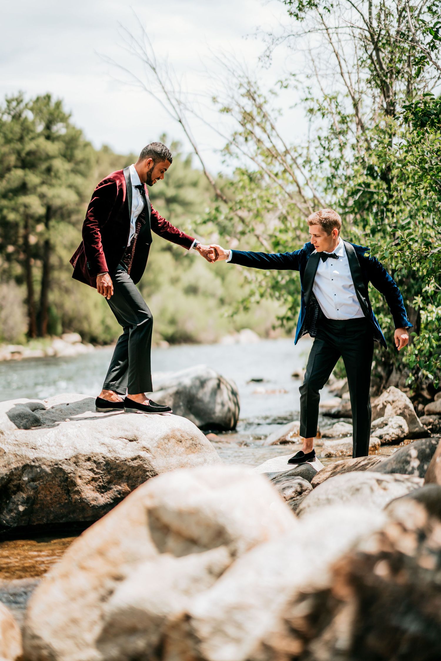 A couple in tuxedos holds hands while balancing on rocks near a scenic river.