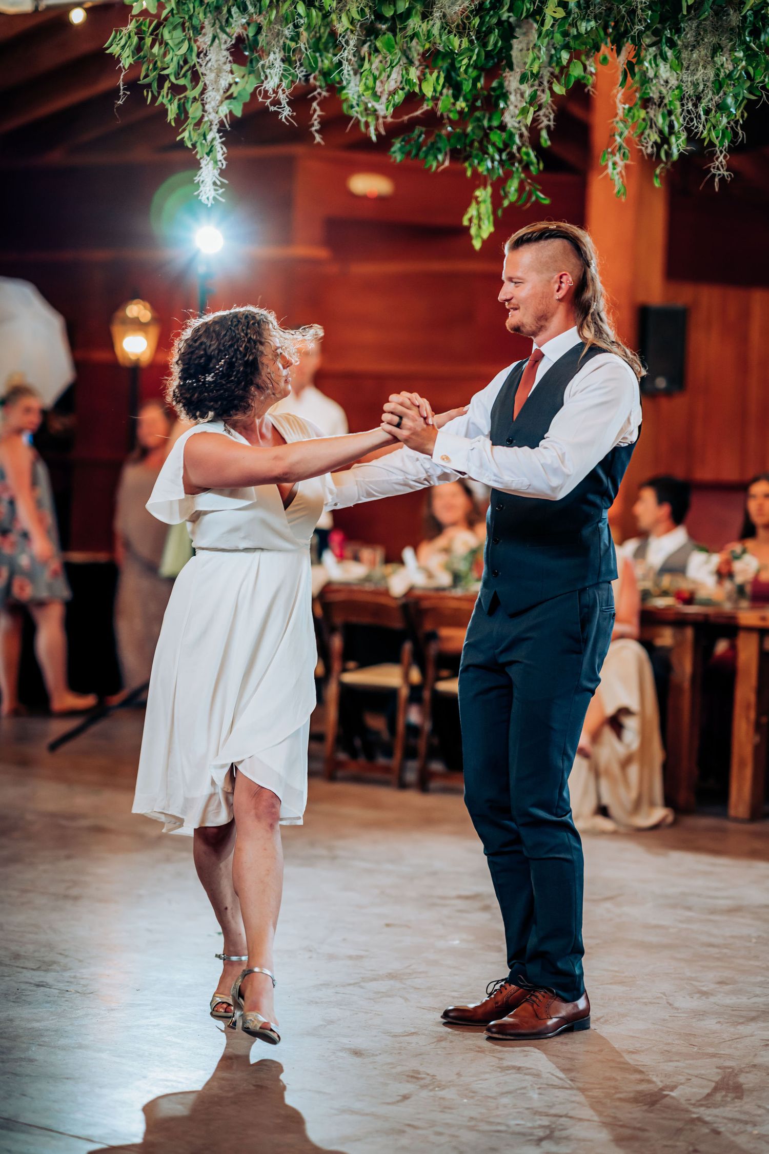 Dancing couple in formal attire perform dance moves under decorative greenery and lights.