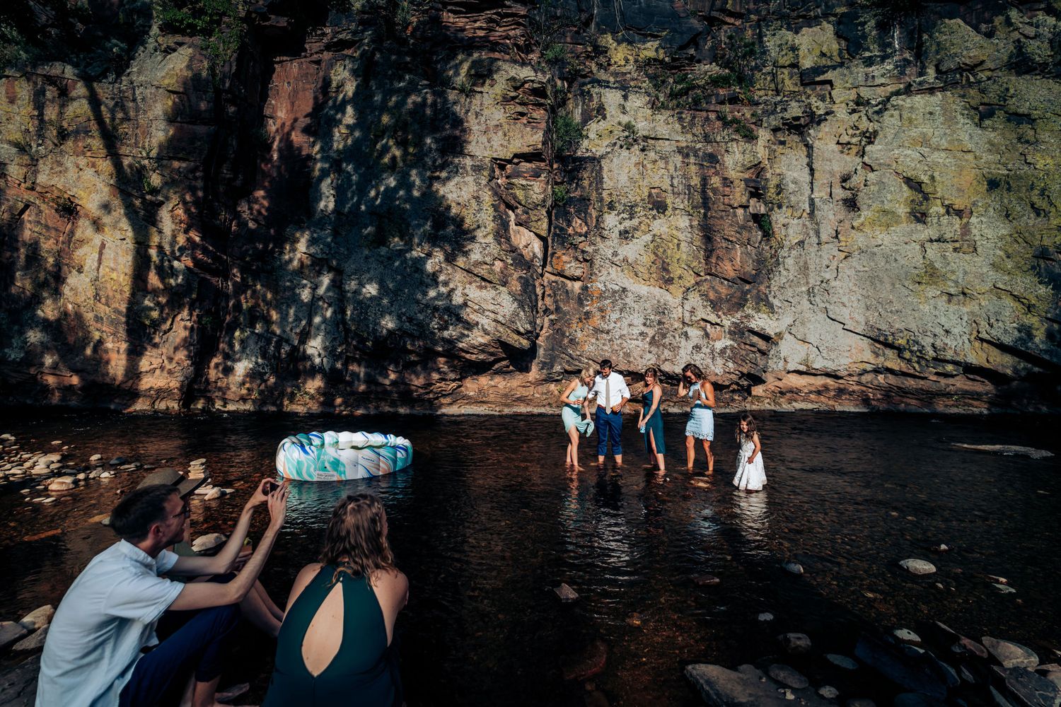Group gathers at the base of a dramatic rock formation near a serene water body.