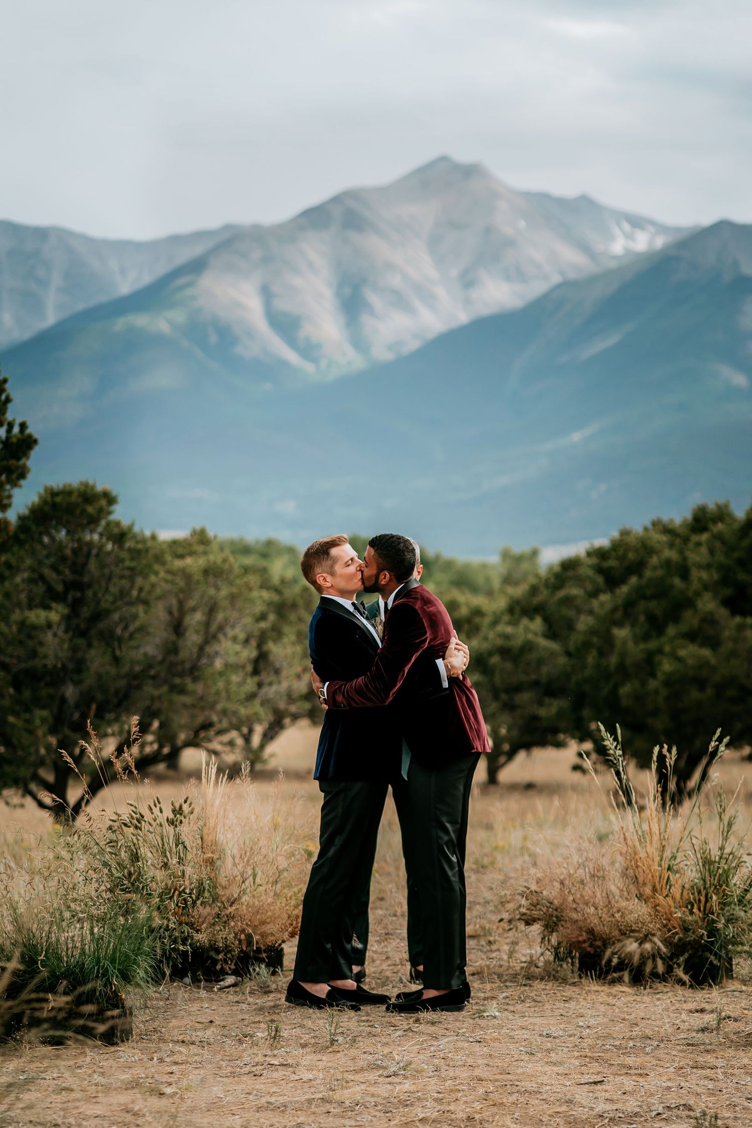 A romantic couple embraces against a stunning mountain backdrop with blue peaks and green foliage.