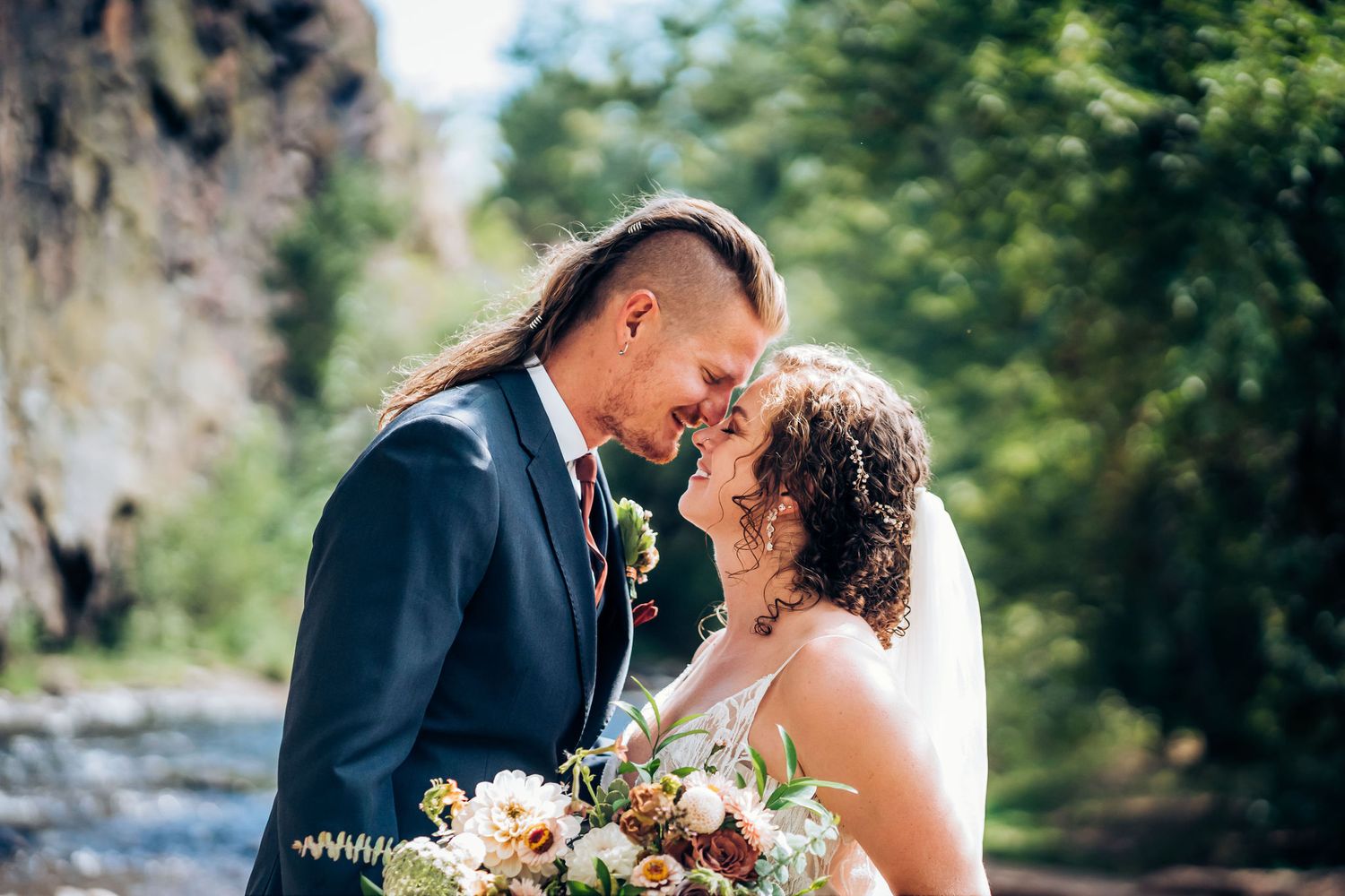 Intimate wedding portrait with blooming bouquet against natural river and cliff backdrop.