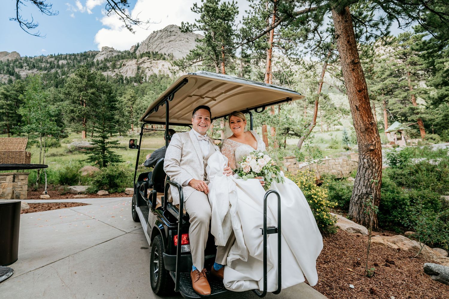 Newlyweds ride together in golf cart through mountain resort setting.