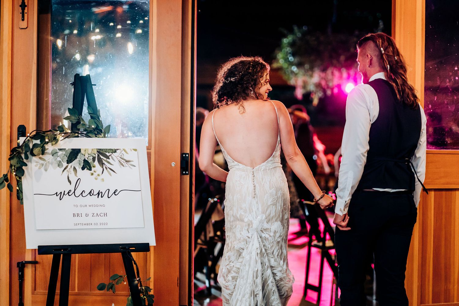 Newlyweds hold hands as they walk past a welcome sign at their reception venue.