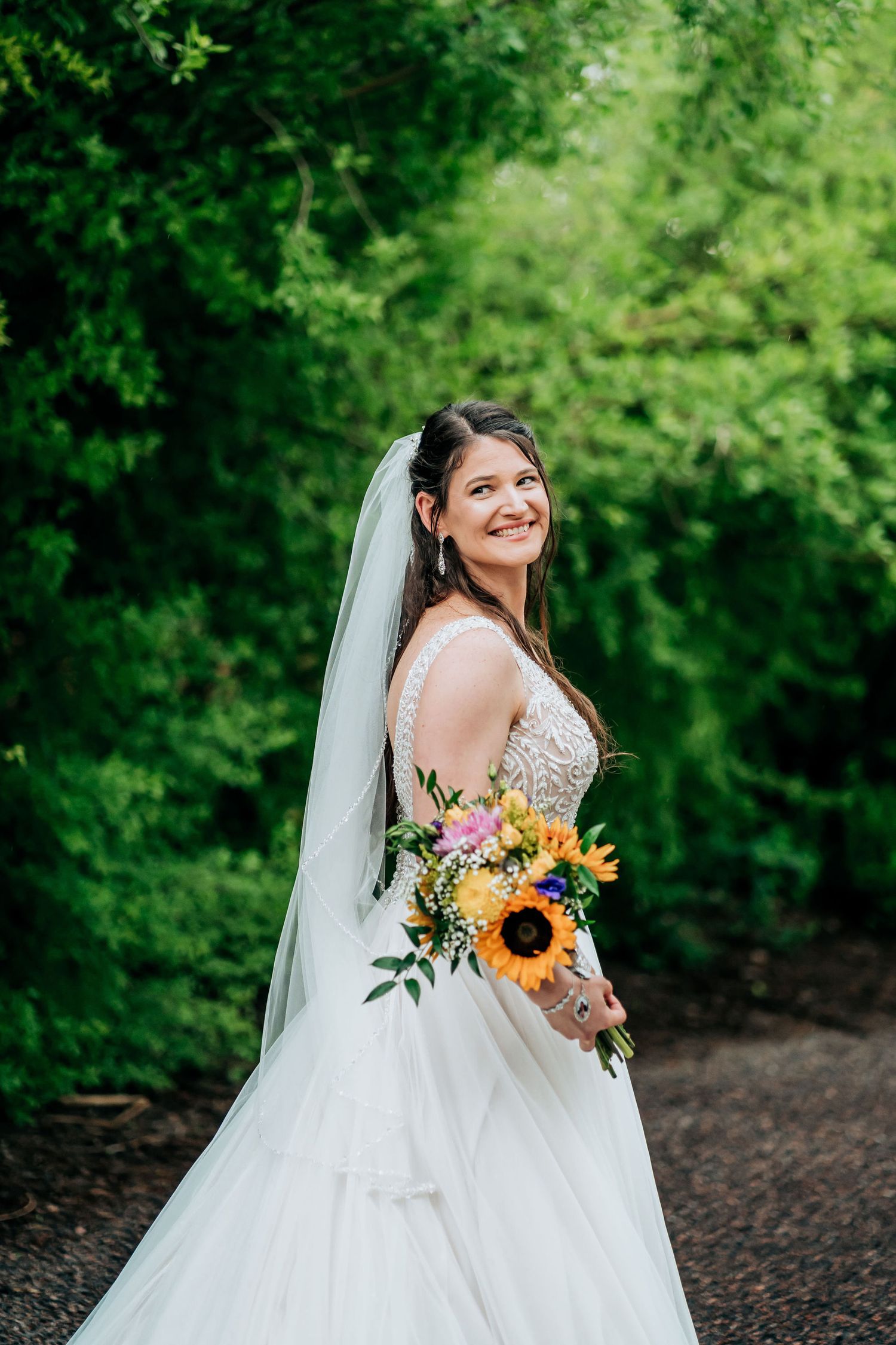 Bride holding vibrant sunflower bouquet poses in white lace gown against green foliage.