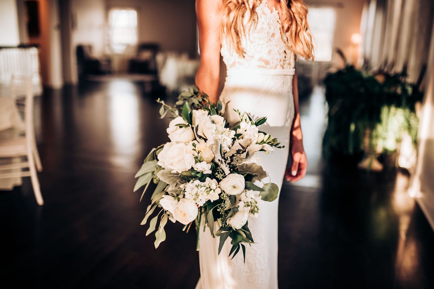 Elegant white wedding bouquet with greenery held against form-fitting bridal gown in indoor setting.