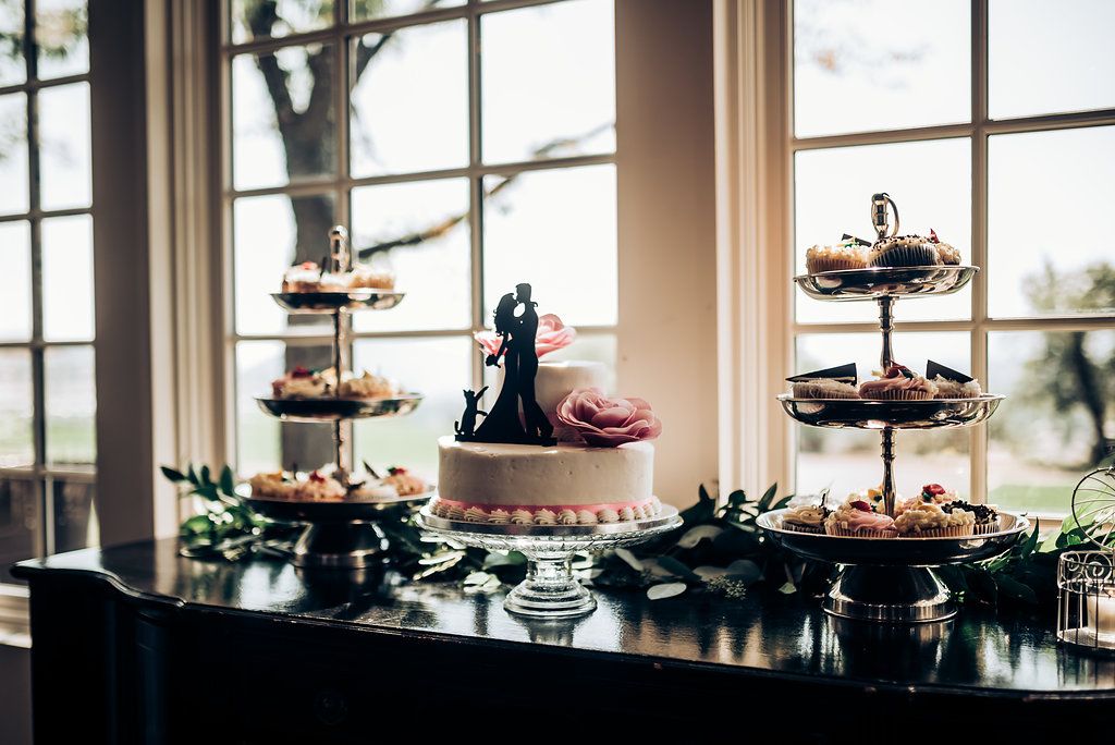 Wedding dessert display with multi-tiered cake and treats on wooden table by window.