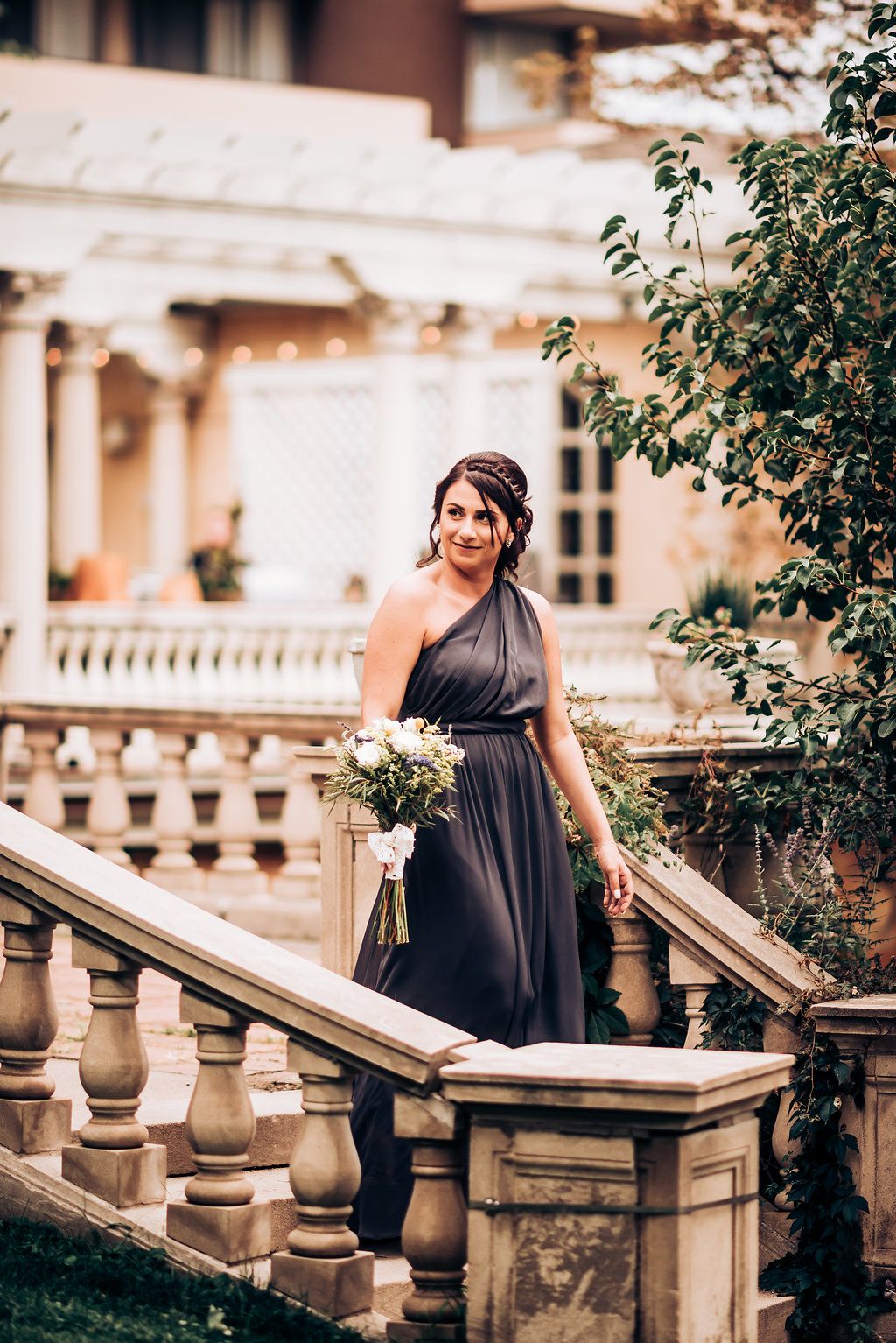 Bridesmaid in dark gown poses on ornate stone staircase holding bouquet.
