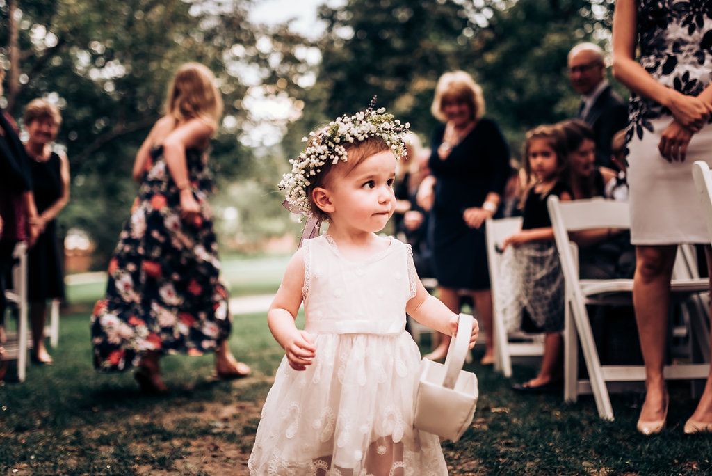 Young flower girl in white dress and floral crown carries basket at outdoor wedding.