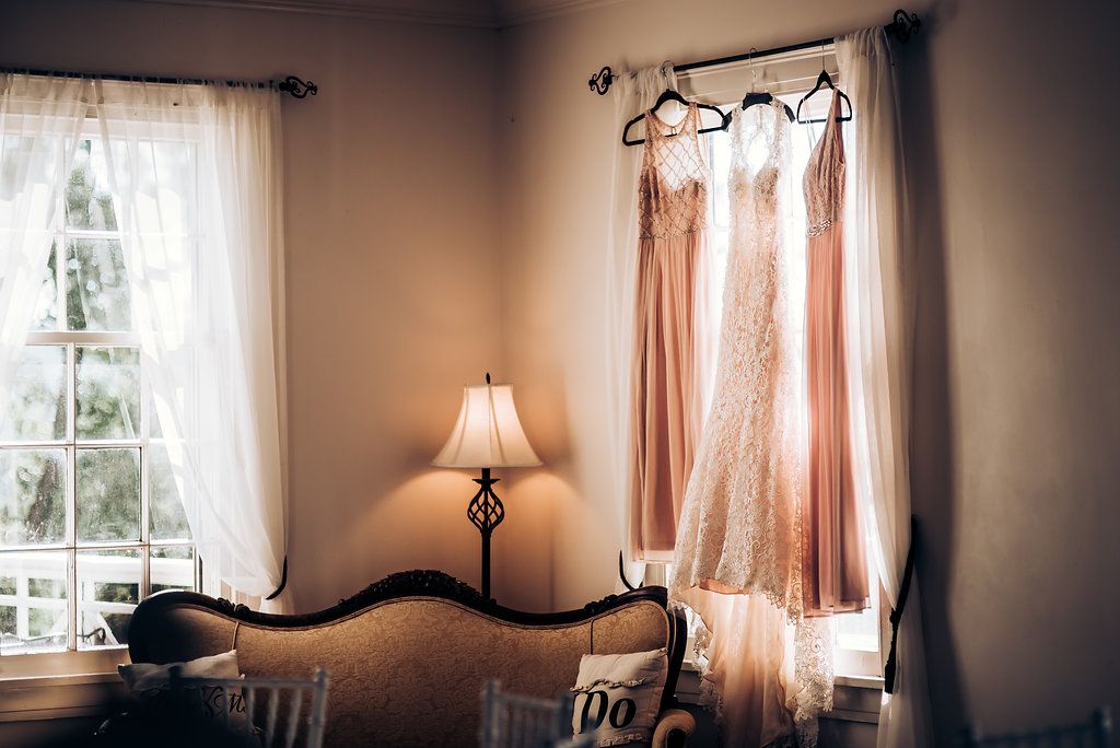 Wedding dresses in blush tones hanging by window with vintage table lamp setting.