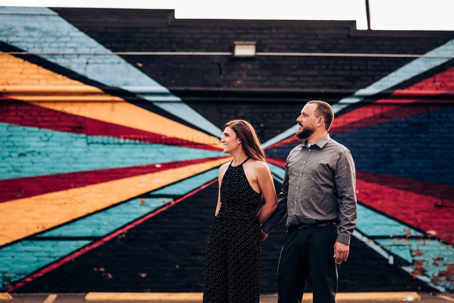 A couple poses against a vibrant geometric mural featuring colorful triangular shapes on a dark wall.