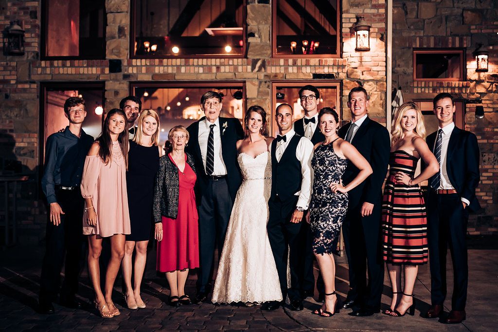 Group photo of wedding party members in formal attire standing in rustic indoor venue.
