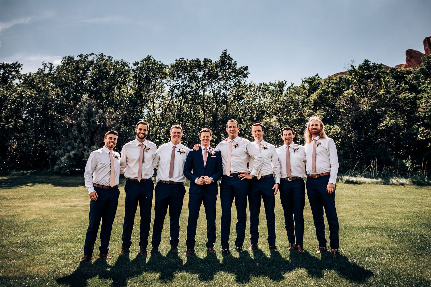 Wedding party groomsmen in matching blue suits posing outdoors on a sunny day.