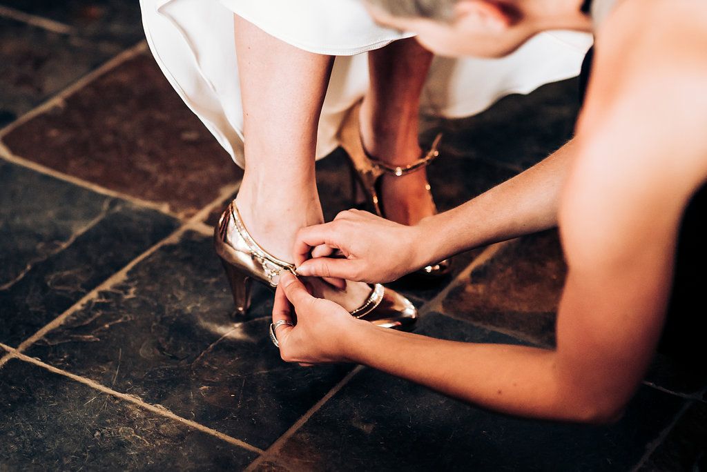 Hands fastening straps on white heels against dark wooden floor.