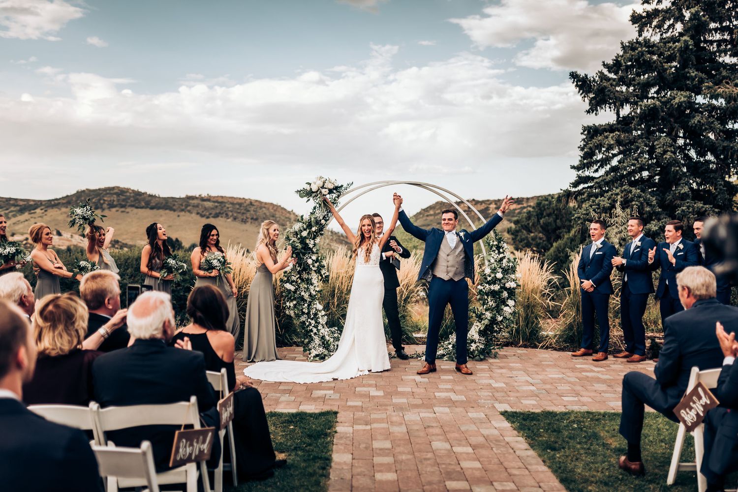 Outdoor wedding ceremony with guests seated on white chairs as couple celebrates under mountain landscape.