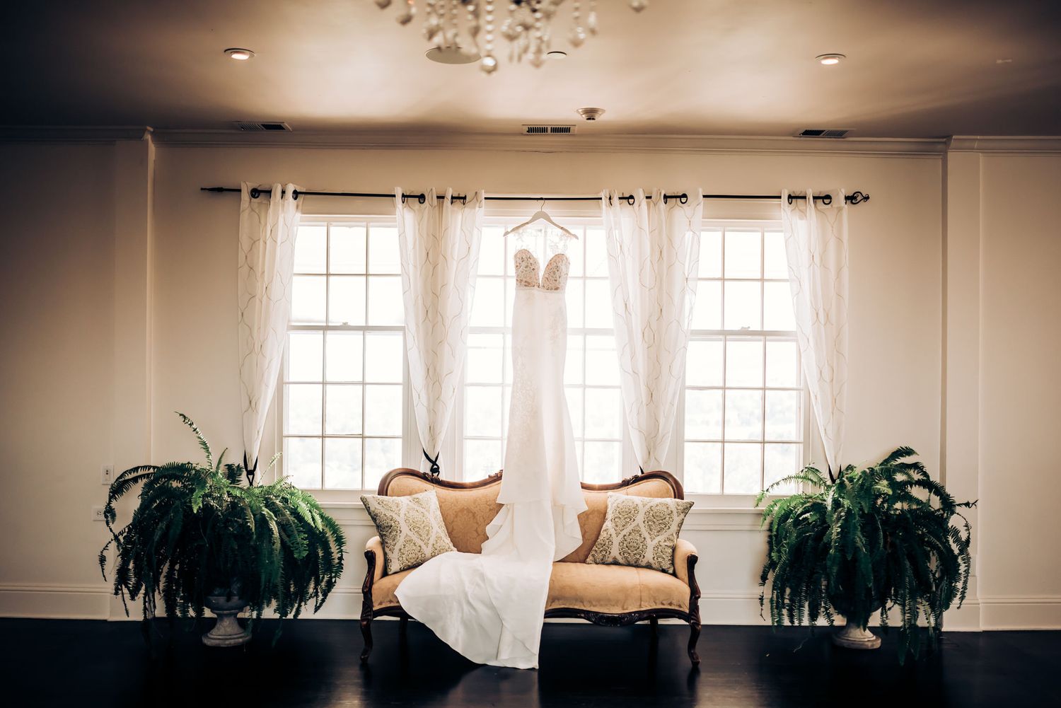 An elegant interior room with three windows, potted plants and a vintage settee draped with fabric.