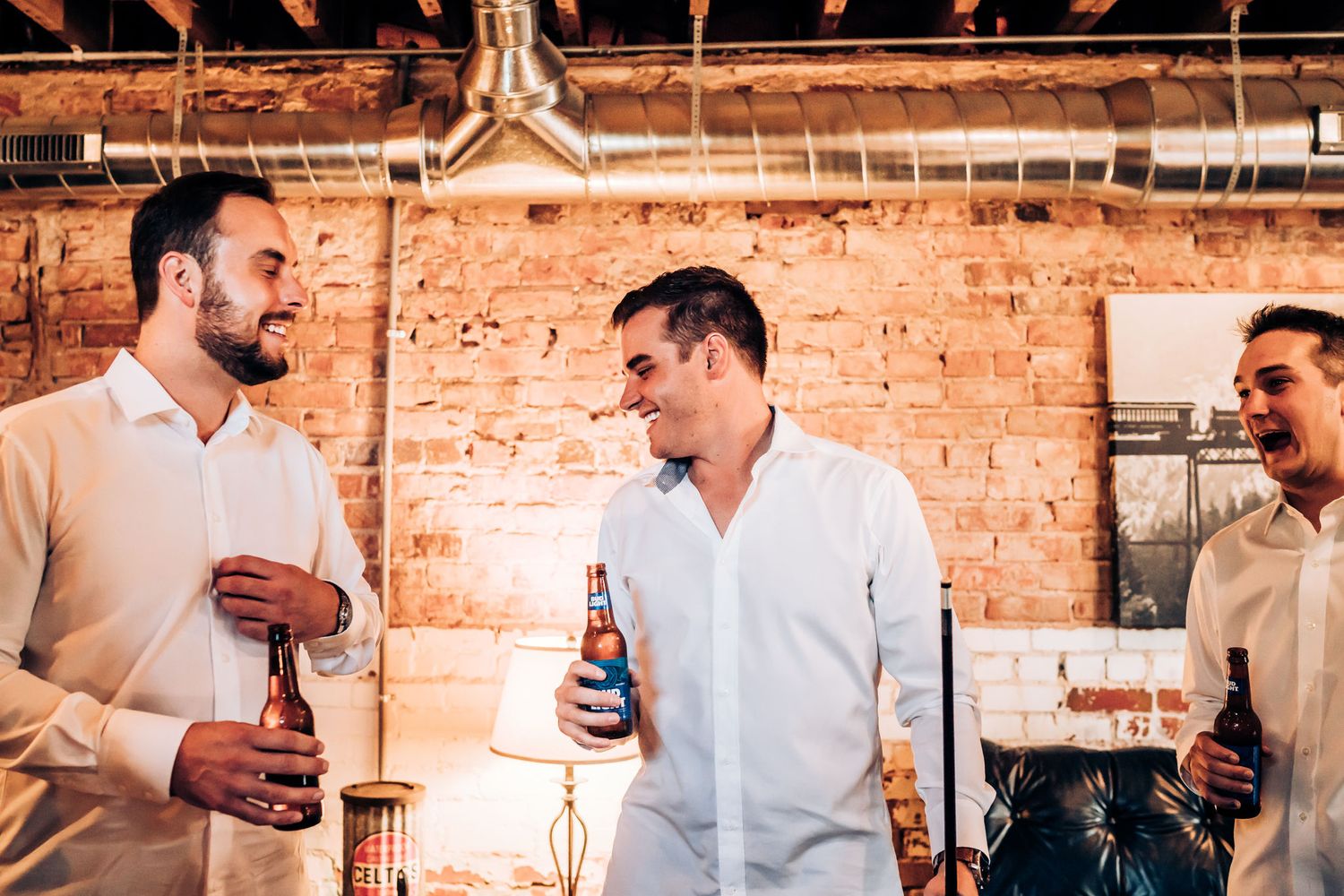 Three men in white shirts laughing and drinking beers at indoor party with exposed brick wall.
