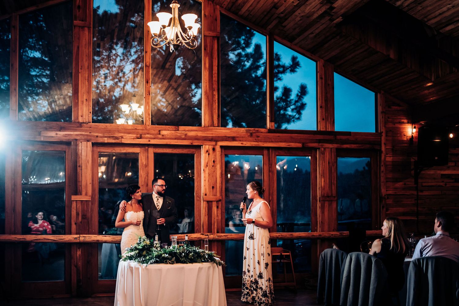 Evening wedding reception in lodge with dramatic floor-to-ceiling windows and wooden beams.