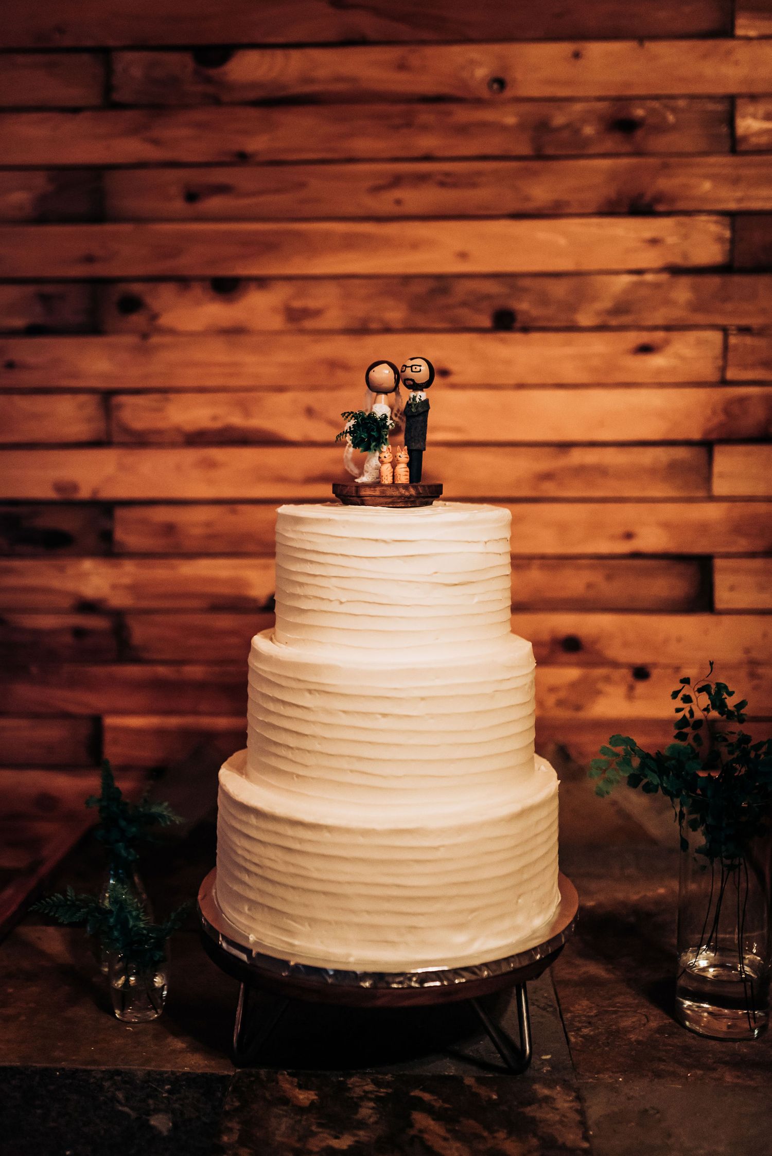 Three-tiered white buttercream wedding cake with custom topper against rustic wooden wall background.