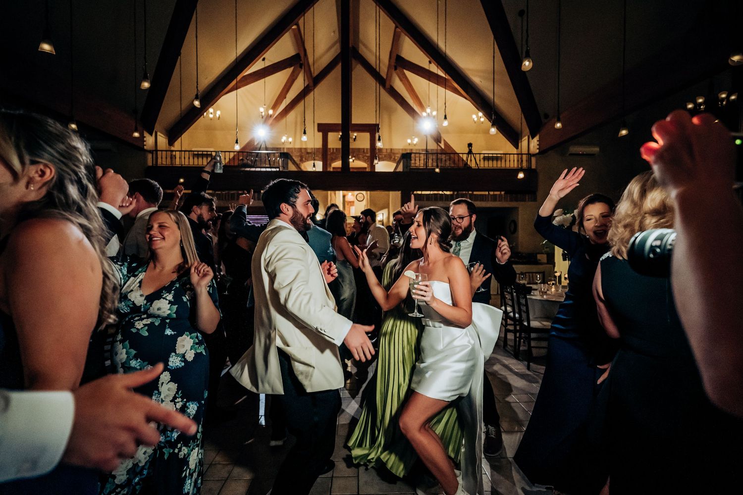 Wedding reception dance party in full swing under vaulted wooden ceiling with dramatic lighting.