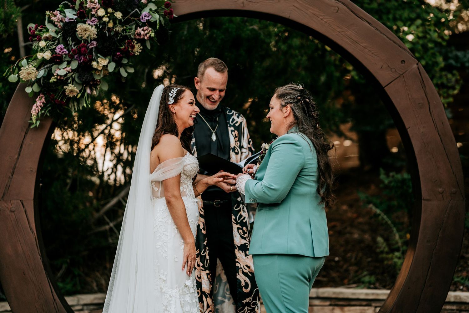 Two brides laugh joyfully with officiant during ring exchange under floral circular arch outdoors.