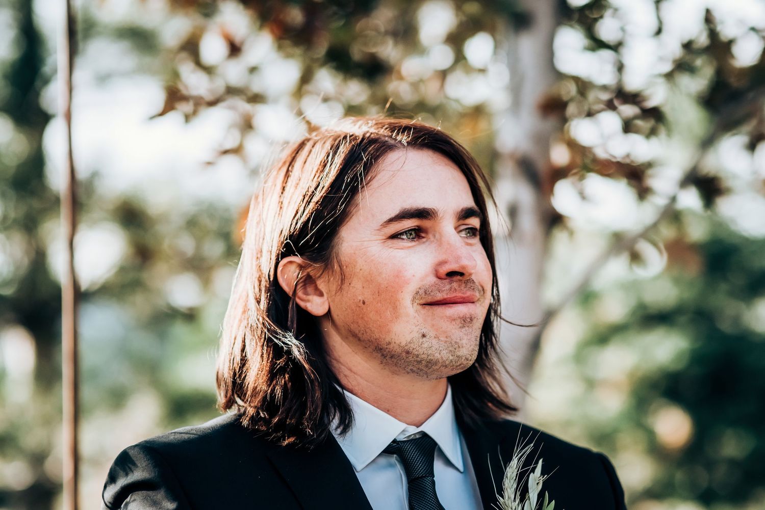 Groom in formal attire with long hair and tie stands in natural outdoor setting.