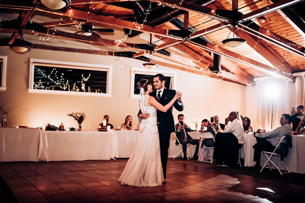 Couple shares first dance on wooden floor under exposed beam ceiling with string lights.