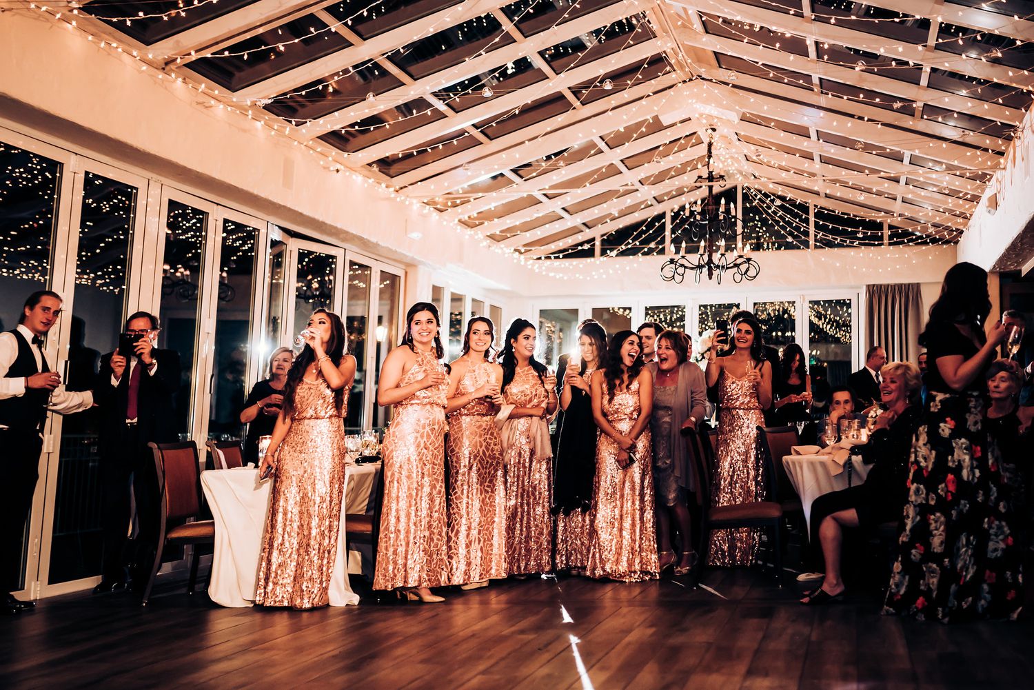 Bridesmaids in shimmering rose gold dresses stand in line at wedding reception.