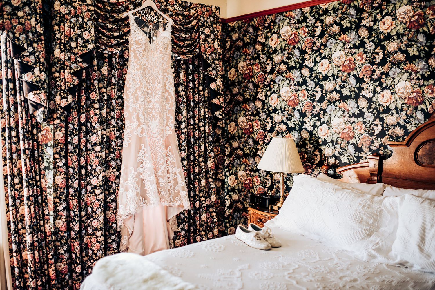 Wedding dress hanging in a bedroom with matching floral wallpaper and white bedding.