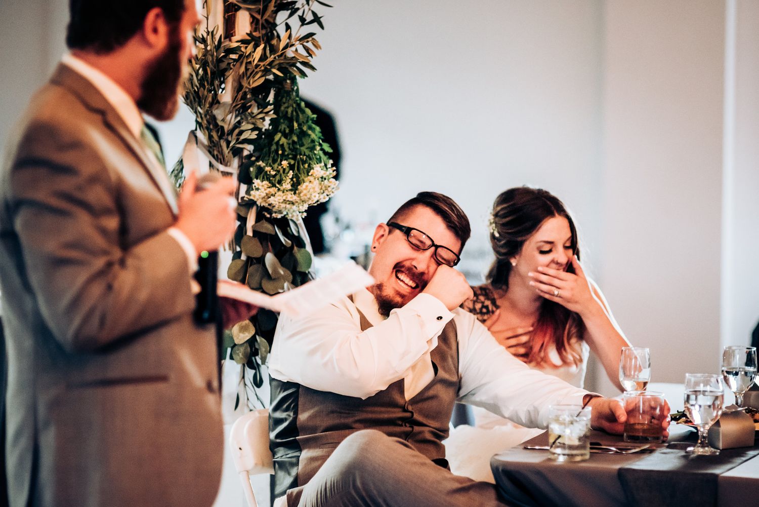 Wedding guests react with laughter during a toast at an indoor reception with greenery decor.