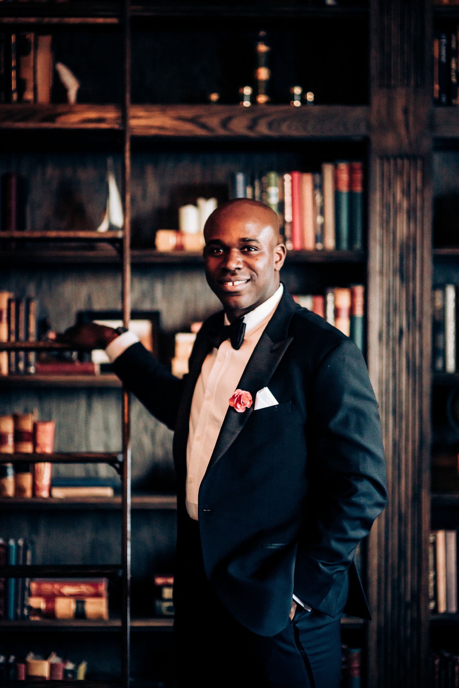 Groom in black tuxedo poses confidently in front of library shelves.