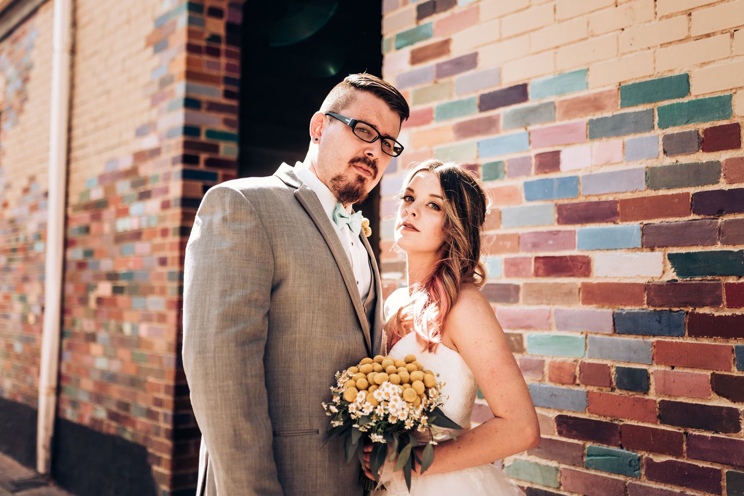 A wedding portrait against a multicolored brick wall with a yellow bouquet.