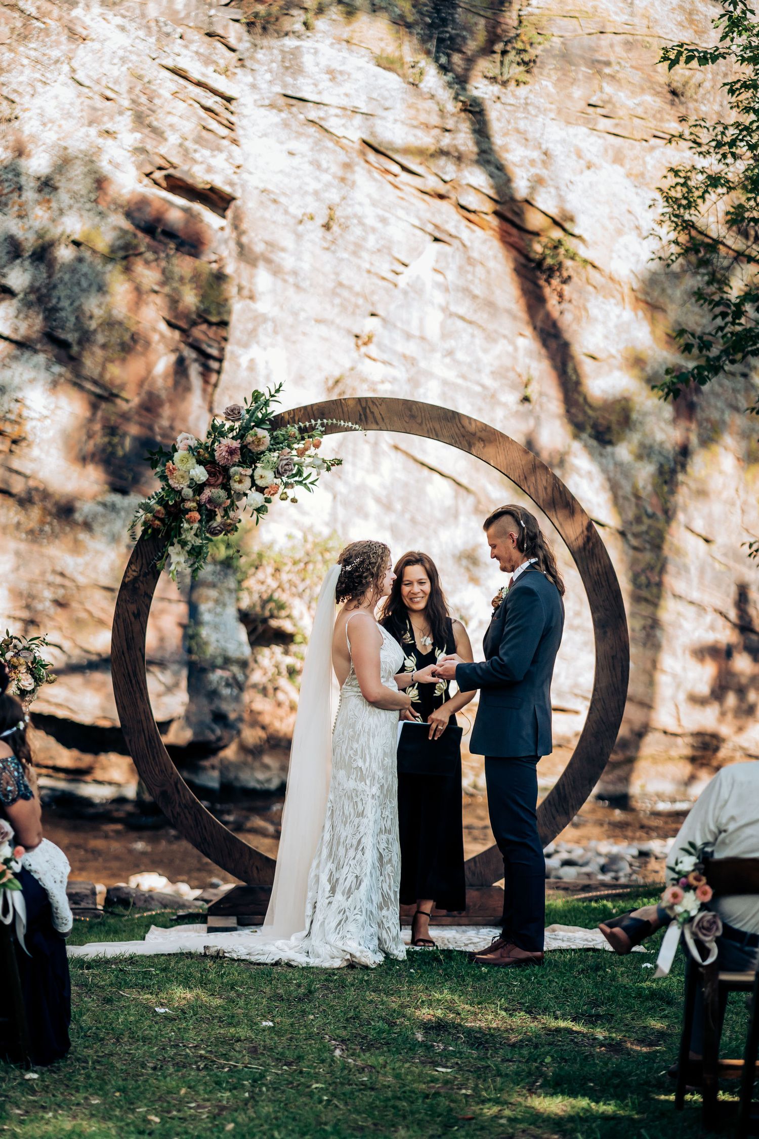 Intimate outdoor wedding ceremony with circular arch backdrop against dramatic stone cliff wall.