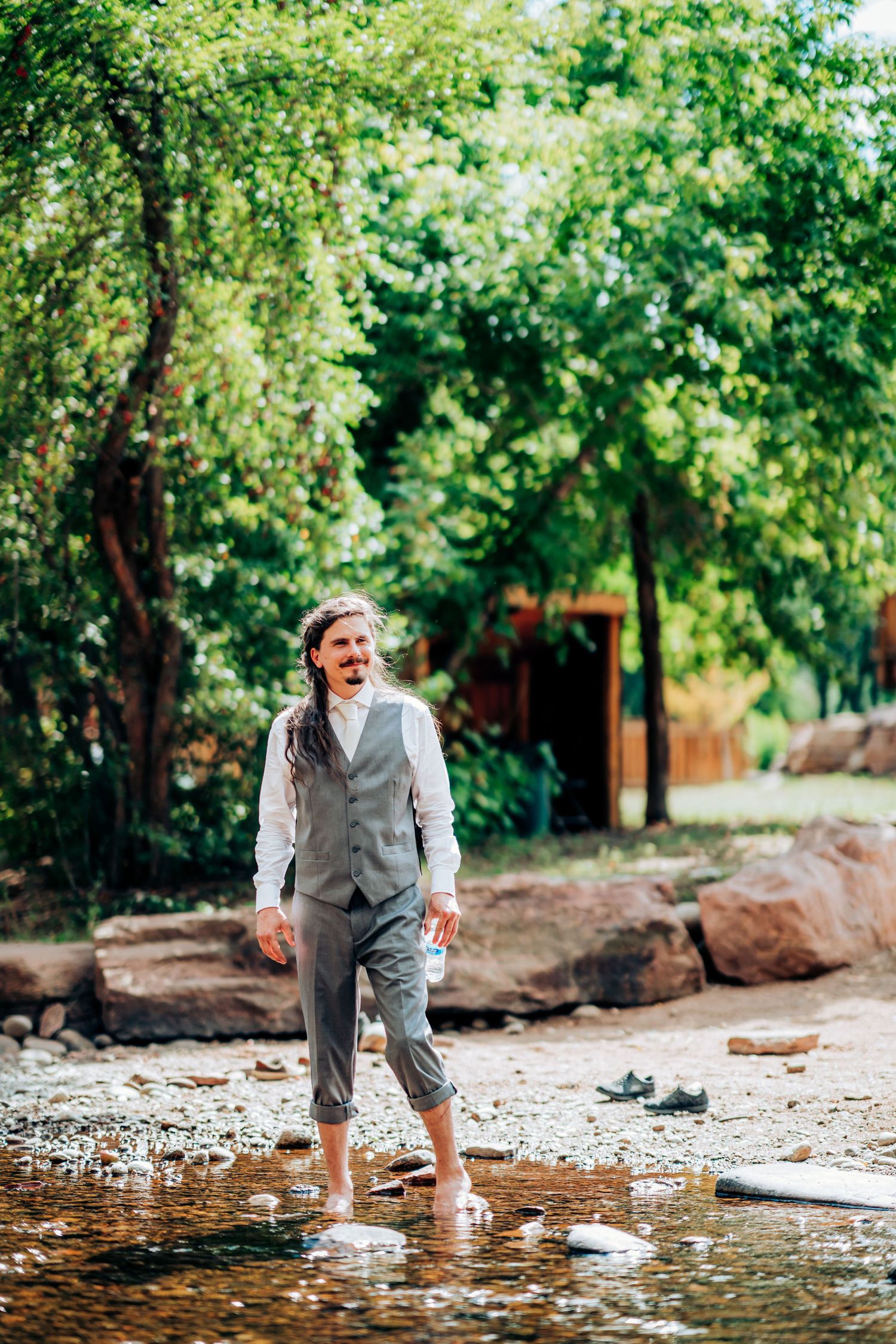 Wedding party member stands in shallow stream wearing grey vest and rolled pants.
