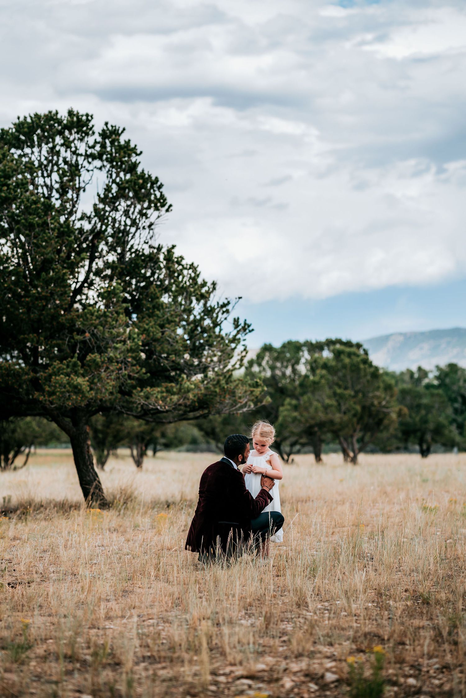 Person in formal attire kneeling in field holding small child with mountains in background.