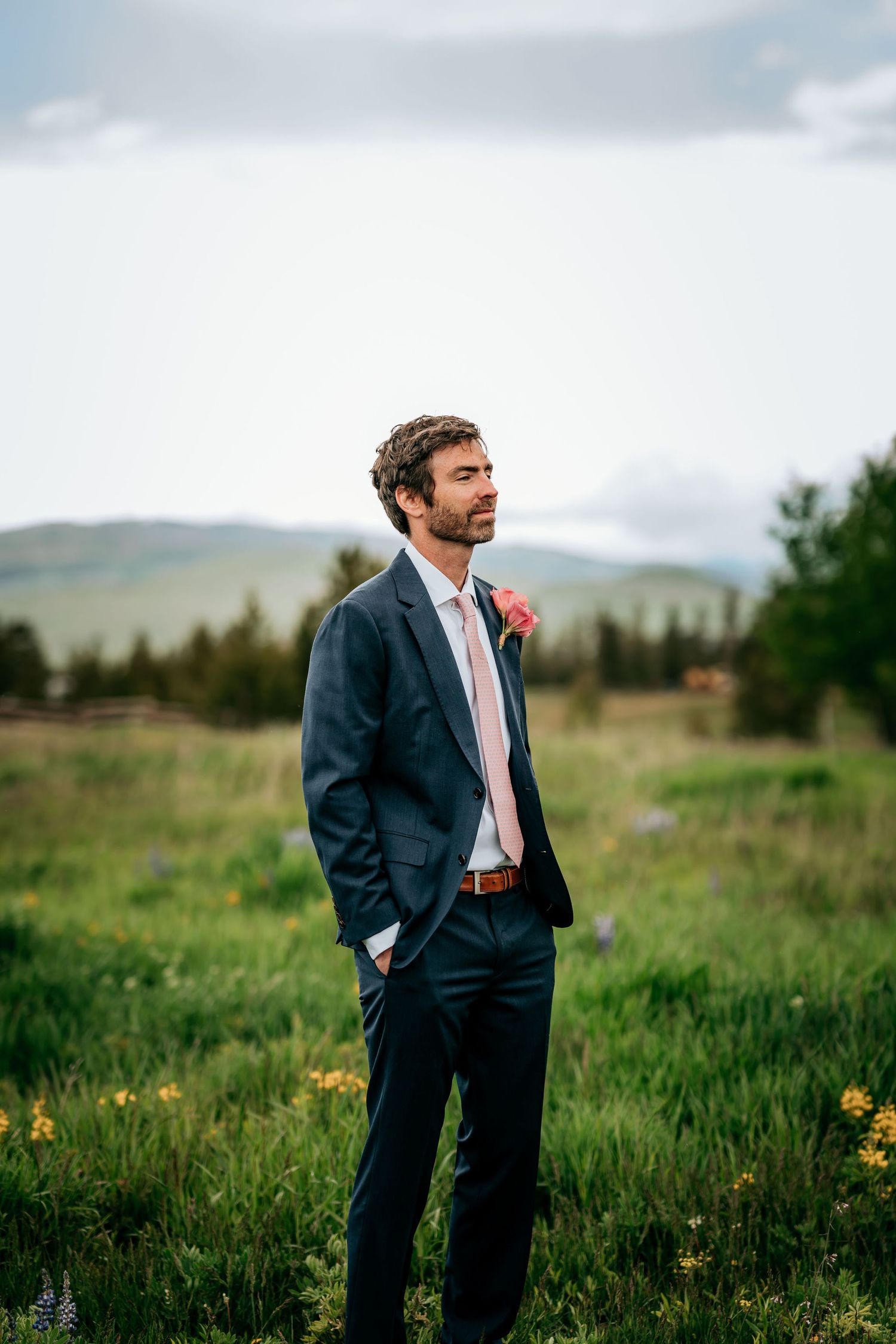 A formal portrait of a suited figure standing in mountain meadow with pink boutonniere.