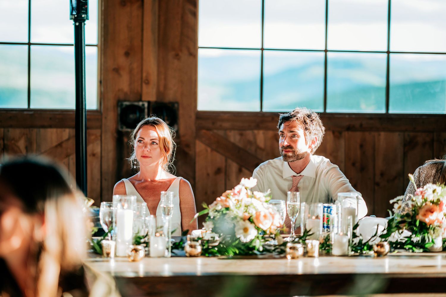 Guests seated at an elegant wedding reception table with floral centerpieces near large windows overlooking mountains.