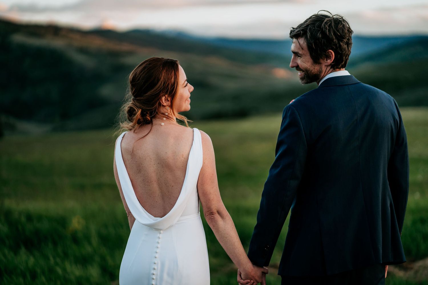 Elegant backless wedding dress detail as couple walks through mountain meadow.