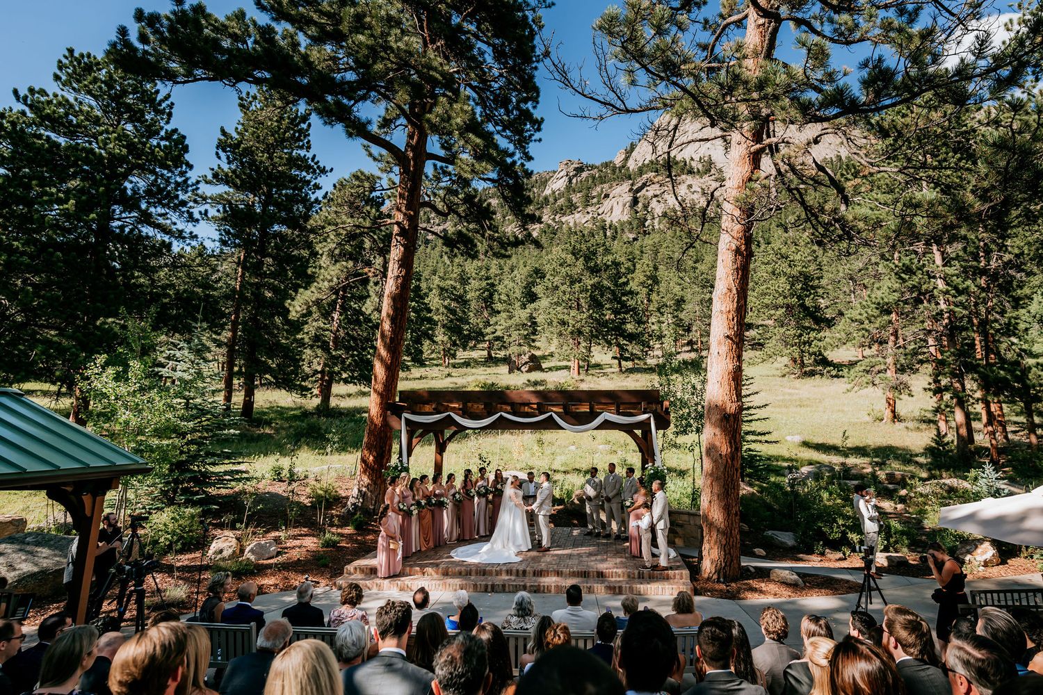 An outdoor mountain wedding ceremony with wooden arch and guests seated among tall pine trees.