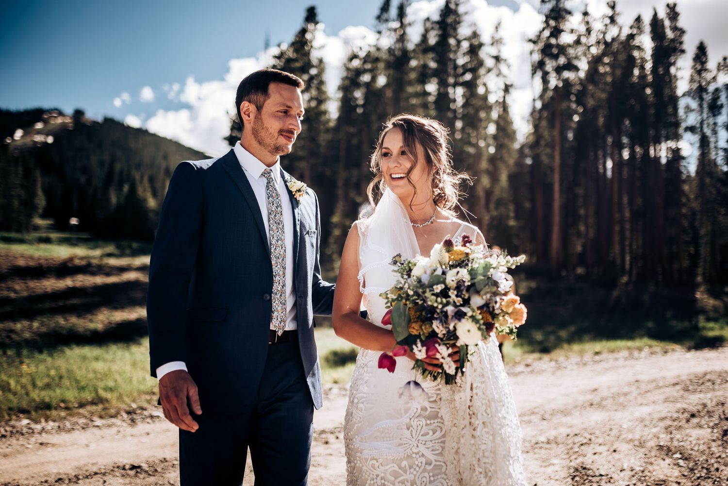 Mountain wedding portrait with bride holding bouquet in lace dress beside groom in navy suit.