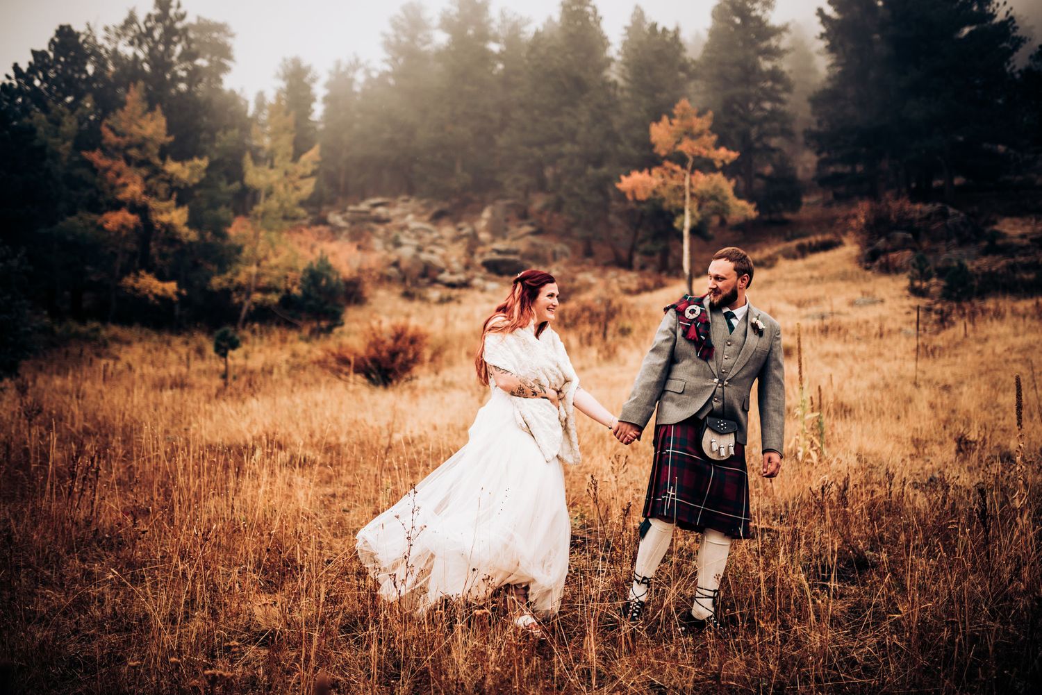 Newlyweds walking through misty mountain meadow with groom in traditional Scottish kilt.