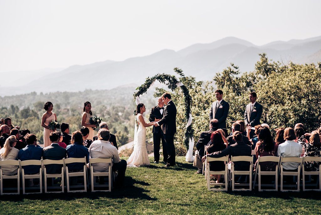 Outdoor mountain wedding ceremony with floral arch and wooden chairs on grassy hillside.