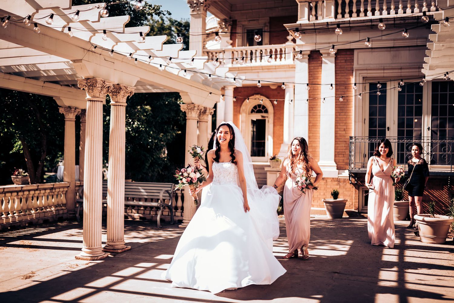 A bride and bridesmaids in blush dresses walking along an elegant covered porch.