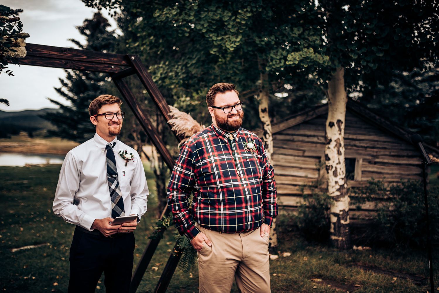 Two people in plaid shirts pose outdoors with rustic cabin in background.