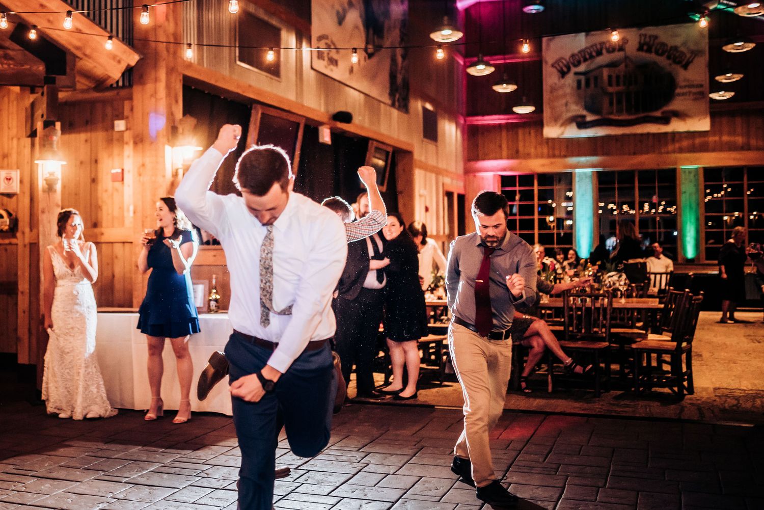 Party guests dance energetically on wooden dance floor under warm venue lighting.