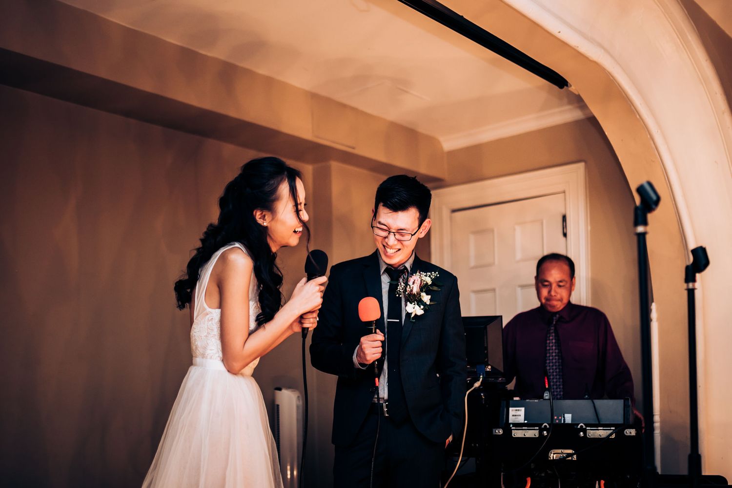 Couple sings karaoke together at a wedding reception while DJ performs in background.