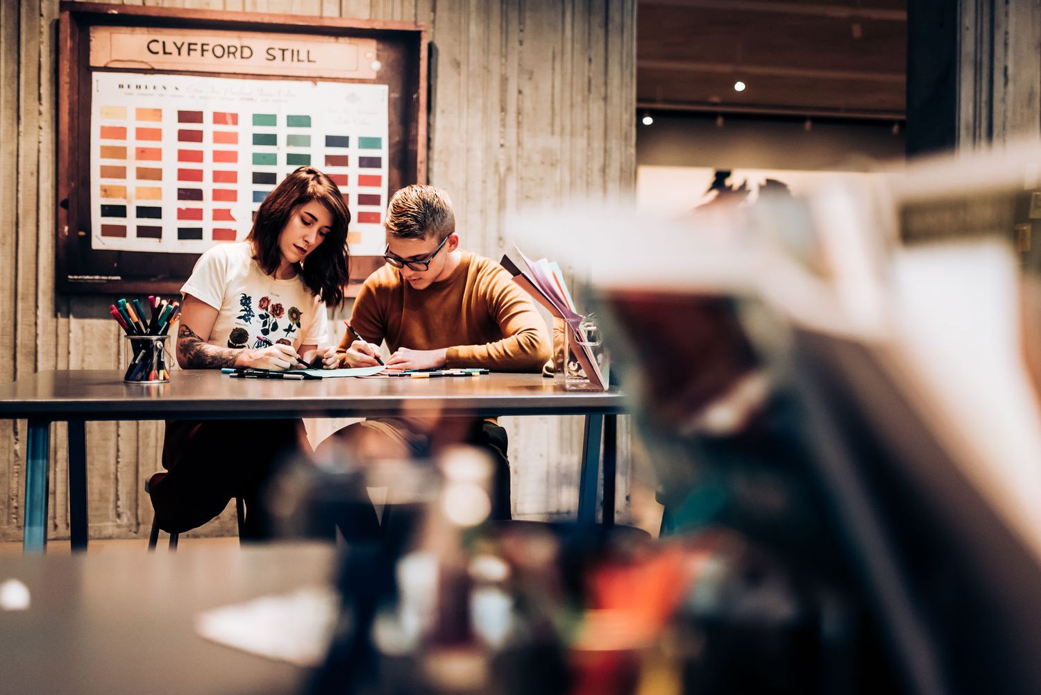 Two people collaborate on artwork at a desk with color swatches displayed on the wall.
