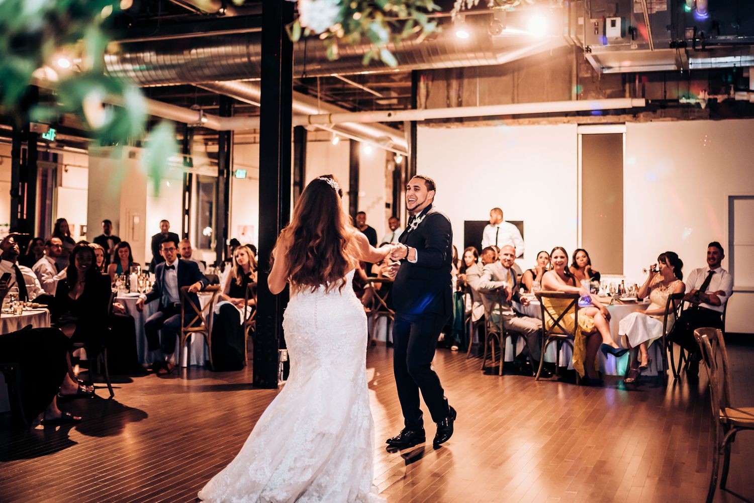 A couple dances together in an industrial-style wedding venue with exposed ceilings and warm lighting.