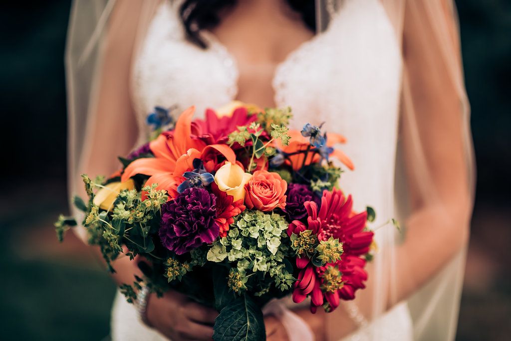Vibrant wedding bouquet featuring coral, pink, and purple blooms with lush greenery against a dark background.