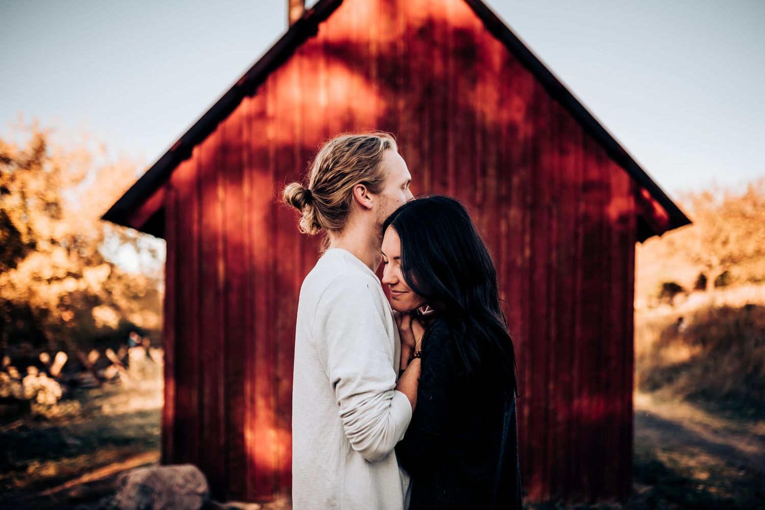 A couple stands close together in front of a red barn during golden hour.