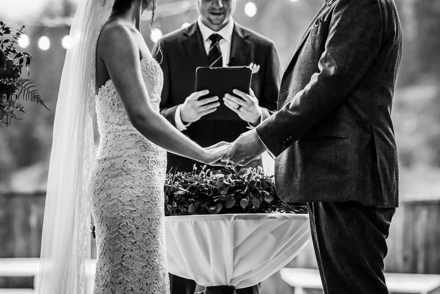 A black and white photo capturing a wedding ceremony with the couple holding hands and officiant reading from a book.